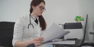 一名戴眼镜的年轻女医生通过视频通话技术与患者进行虚拟会面，她手里拿着患者的测试结果，并在诊所办公室的笔记本电脑上给出治疗建议