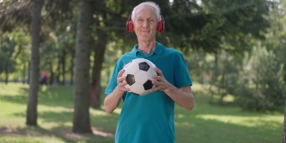 戴着耳机的快乐老人在夏日公园里与足球摆姿势。积极的白人男性退休人员看着相机微笑享受春天户外运动的肖像。长寿的概念。