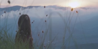 一个年轻女子坐在草地上的后视图，看着从山上落下的厚厚的白色雾