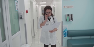 医院的医生一边走在现代诊所的走廊上一边翻阅病历，一边打电话。女医生在医院走廊里用手机阅读文件