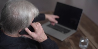 从肩膀上看到的无法辨认的白发苍苍的老年男性与技术支持人员交谈，请求帮助修理坏了的笔记本电脑。