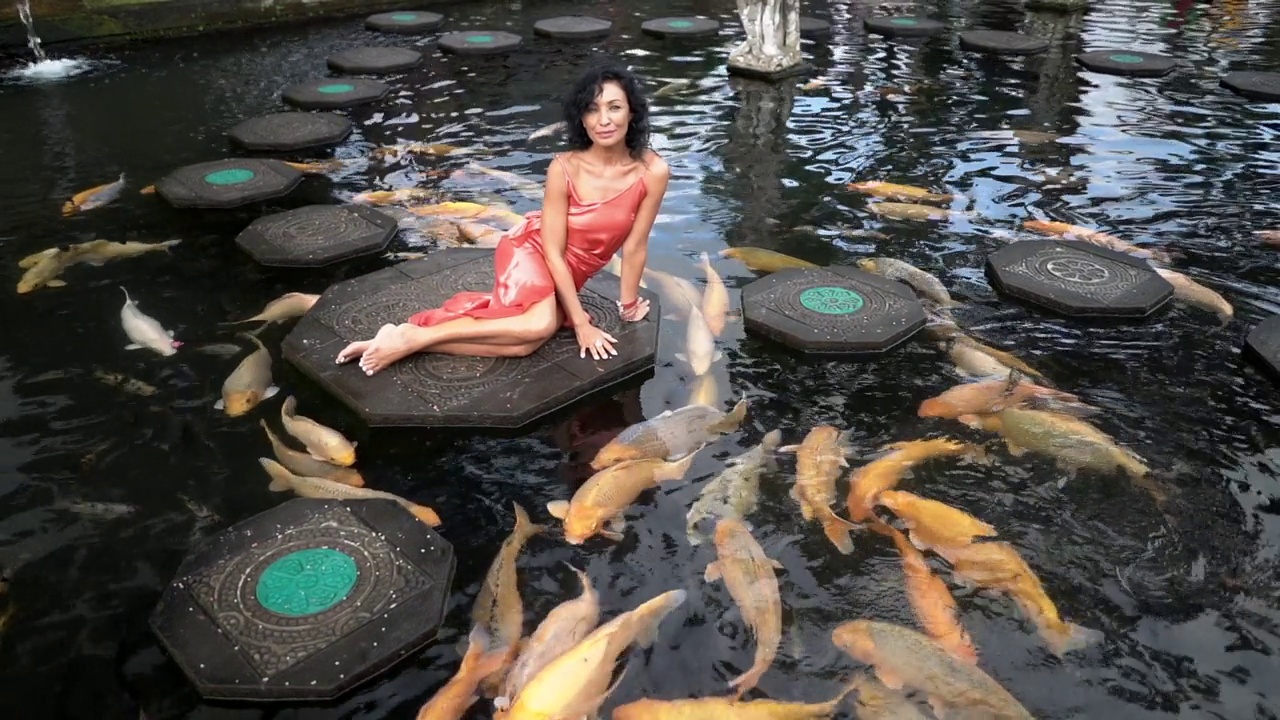 迷人的深色皮肤的模特穿着红色的衣服，放松在石径上的池塘五颜六色的鱼
