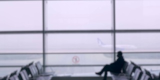散焦的机场背景，一名男子坐在登机口，窗户上似乎有一架商用飞机正在跑道上。男人正在看他的手表。
