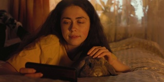 一个微笑的女人趴在床上抚摸着一只垂耳兔，一边看着手机。宠物护理。家庭娱乐