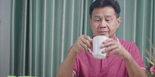 亚洲老男人从白色的杯子喝热饮，休息一天坐在家里的办公桌，退休生活，成熟的人类阶段，早上咖啡咖啡开始新的一天