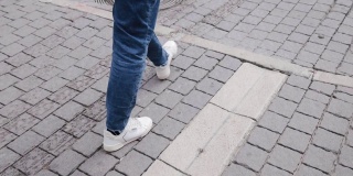 这是一个穿着白色运动鞋和牛仔裤的男子的腿部特写镜头，他自信地走在特拉维夫街头的人行道上。50帧/秒慢动作
