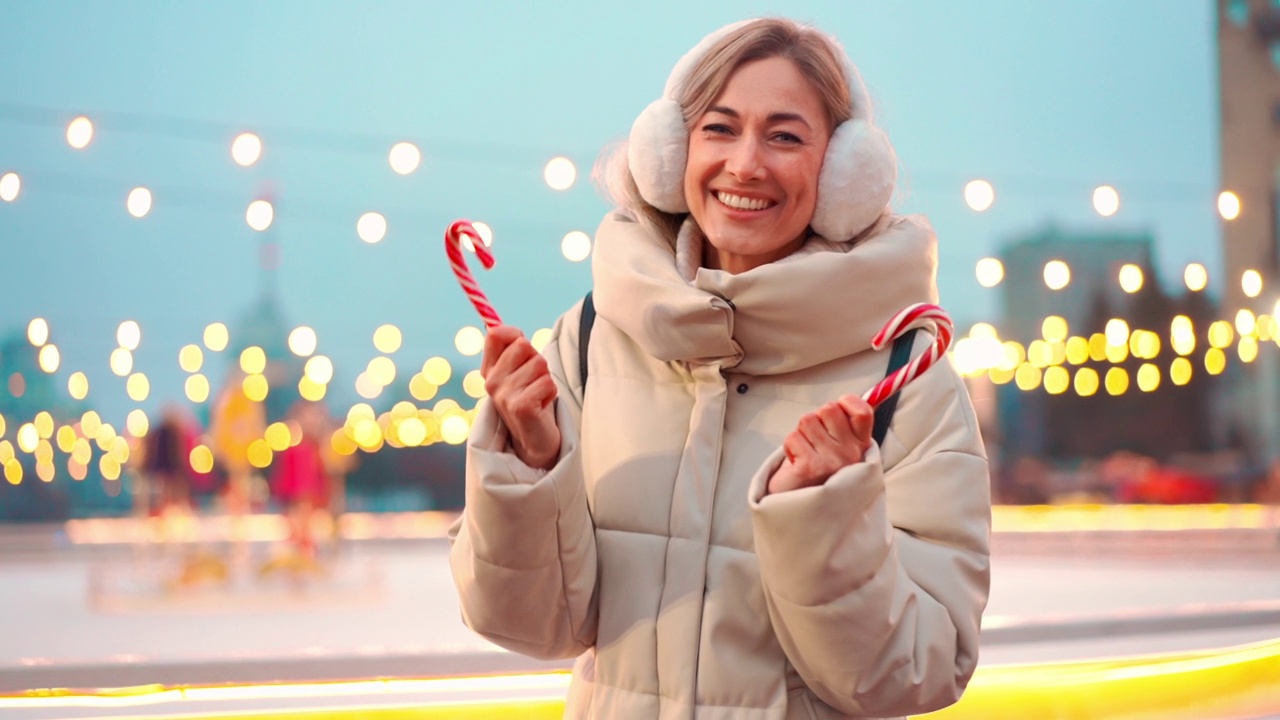 冬季假期晚上，一名女子戴着耳套站在市中心广场的溜冰场附近。人们在圣诞灯的背景下滑冰。