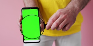 一名身穿白色牛仔裤和黄色t恤的男子展示了一个绿色智能手机屏幕，并将放大镜移到屏幕上。模拟。粉色背景,特写。搜索和广告的概念