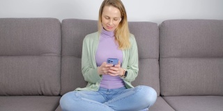 一名悠闲的白人女子，盘腿坐在家里的沙发上，用智能手机打字，戴着无线耳机听音乐。美丽的女性用手机在线交流