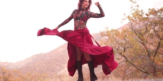 华丽的红发魔术女人在一个红色的长裙在山的金色森林的边缘跳舞