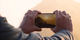 男性的手用智能手机和点击Girnar山的照片的特写。摄影师创造多媒体内容的山在日出，男子点击图片在徒步冒险。