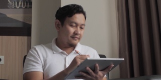 一名亚洲男子坐在客厅的沙发上，用手写笔在他的数字平板电脑上画画，这位年轻的数字企业家喜欢在家工作
