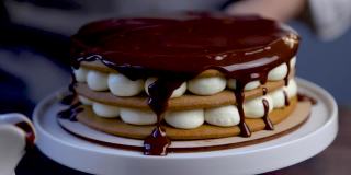 蛋糕有三层，蛋糕和白奶油放在一个架子上，上面装饰着白奶油和融化的巧克力，厨师用铁铲把巧克力撒在蛋糕表面，巧克力顺着边缘流下来