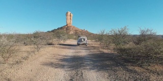 无人机航拍的视频片段，一辆白色4x4驱动吉普车在非洲纳米比亚向令人印象深刻的岩石纪念碑Vingerklip行进。在非洲沙漠中形成的高大的岩石。无人机跟随白色货车探索非洲内陆。