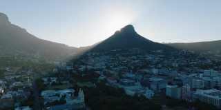 在住宅区高高的山顶后面升起的太阳。狮头山投下的影子在小镇上。南非开普敦