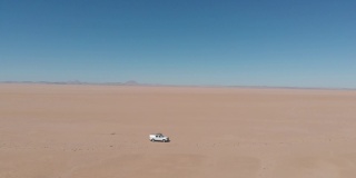 无人机航拍的视频画面显示，一辆白色四驱车吉普车驶过非洲纳米比亚的沙漠。一辆白色轿车孤立地开向远方。越野和超载的背包探险旅行。无人机跟随吉普车探索沙漠。