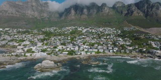 在高高的岩石山脊下的山坡上的自治市的房屋。海浪撞击海岸岩石的向后揭示。南非开普敦