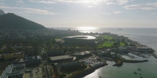 鸟瞰图的海岸和建筑物在海滨。反夕阳的现代圆形足球场结构。南非开普敦