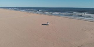 无人机航拍到的白色吉普车在非洲纳米比亚骨架海岸的沙滩上行驶。站在吉普车上看着大海的冒险家和背包客。棕色的沙子和海浪。无人机跟着货车和吉普车。