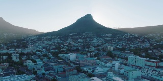 高耸的尖峰在城市的居住区投下阴影。空中上升的镜头显示狮子头山背后的灿烂阳光。南非开普敦