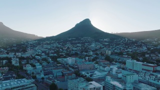 高耸的尖峰在城市的居住区投下阴影。空中上升的镜头显示狮子头山背后的灿烂阳光。南非开普敦视频素材模板下载