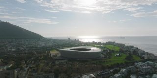 日落时分，在城市的海岸上空向后飞行。绿点体育场现代化结构及周边开发。南非开普敦