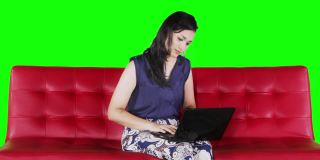 一名年轻女子坐在红色的沙发上，一边思考一边用笔记本电脑，用4k分辨率拍摄，屏幕背景是绿色的