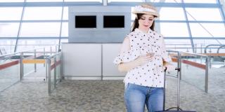 快乐的女性旅客站在机场大厅，用她的手提箱使用手机