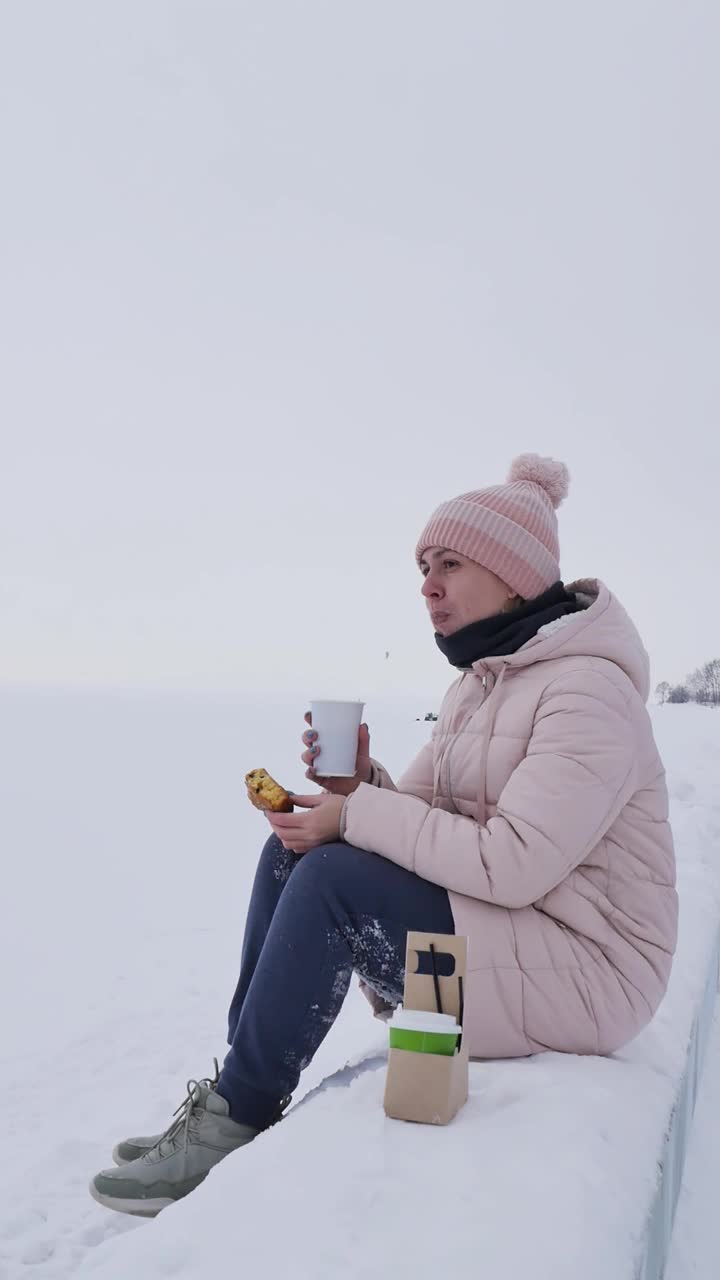 冬天，一位戴着帽子、穿着夹克的年轻女子坐在堤岸的护墙上，吃着杯托蛋糕和卡布奇诺咖啡。冬天冰封的海岸。垂直视频。肖像