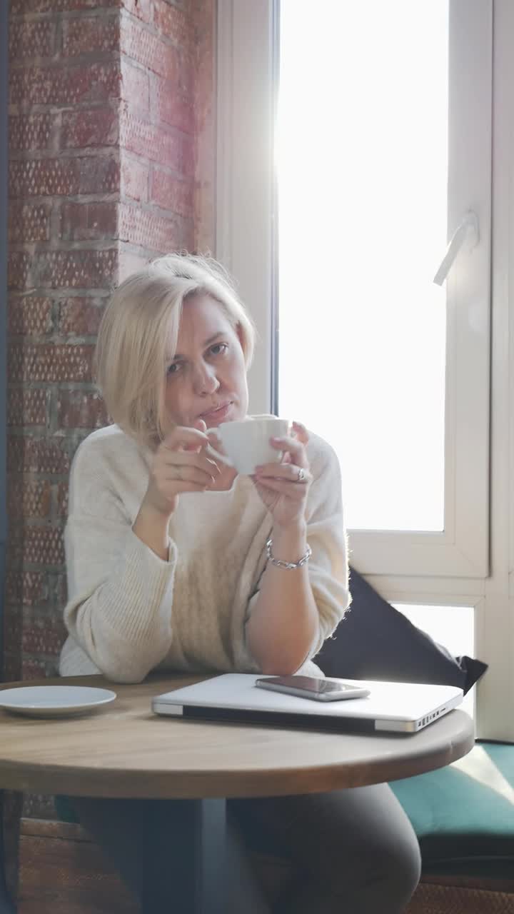 一个美丽的成年金发女郎在靠窗的咖啡馆里拉直头发，用杯子喝着饮料。自由职业者的休息时间。垂直视频
