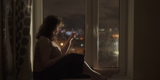 一个女人坐在窗台上，靠近潮湿的窗户。女孩用手指划过玻璃。