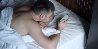 英俊的年轻人在床上打手机。一名白人男子躺在床上用手机上网发短信