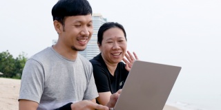一个留着胡子的女人和一个男人坐在沙滩上，手里拿着笔记本电脑。与朋友视频通话，并在一个明亮的日子在海边度假放松