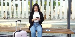 近距离拍摄的好看的积极梦幻的亚洲女孩，穿着休闲的衣服，坐在公共车站的手提箱和打字的电话信息