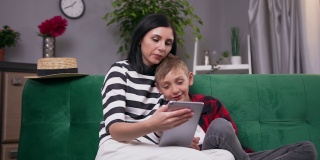 可爱的满意的快乐的现代母亲和儿子一起休息在家里的软沙发上，看有趣的应用程序在i-pad上