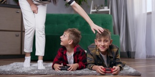 美丽快乐的年轻女子路过她英俊积极微笑的儿子们，他们正躺在地板上用操纵杆玩电子游戏