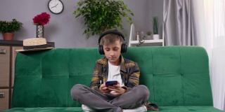 10岁帅气男孩的肖像，戴着耳机，坐在家里的沙发上，一边玩手机一边听音乐