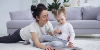 迷人的肖像，快乐的微笑灿烂的母亲，与她的可爱有趣的小女儿在地板上笔记本电脑