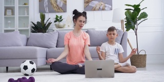 漂亮活泼微笑的年轻女子在地板上的笔记本电脑前和她冷静的十几岁的儿子一起做瑜伽练习