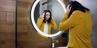 迷人的肖像自信的30岁的黑色头发的商业妇女在时尚的西装站在镜子附近，拉直她的衣服和发型