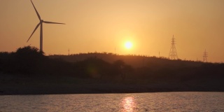 在印度古吉拉特邦的万卡纳，夕阳西下，风车在河边的山丘上拍摄剪影。在黄金时段，除了太阳，还能看到风车和电塔。自然日落背景。