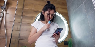 近距离观察满意的漂亮的年轻女子与黑色的头发，站在浴室的镜子和使用她的手机刷牙
