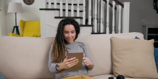 迷人的微笑时尚的年轻孕妇坐在舒适的沙发在客厅和观看喜爱的应用程序在i-pad