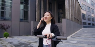 好看的满意的幸运的年轻亚洲女孩在休闲服装享受她的电话交谈，而走在现代建筑附近与自己的电动滑板车