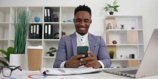 正面看开朗、好看、轻松的年轻黑皮肤男性办公室经理穿着时髦的衣服，在办公室里用手机坐在他的工作场所