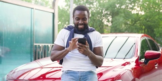 英俊微笑着留着胡子的黑皮肤年轻人站在他自己的红色名车旁洗车，享受着手机上的搞笑照片