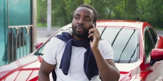 这张照片拍摄的是一位迷人的30岁非洲裔美国人，他面带微笑，留着整洁的胡子，穿着白色t恤，正在洗车时享受着在自己闪亮的红色汽车旁的电话交谈