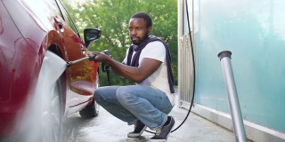 前面的观点，可爱的集中年轻的蓄着胡子的黑皮肤的男人在洗车用电动水洗衣机洗车的车轮