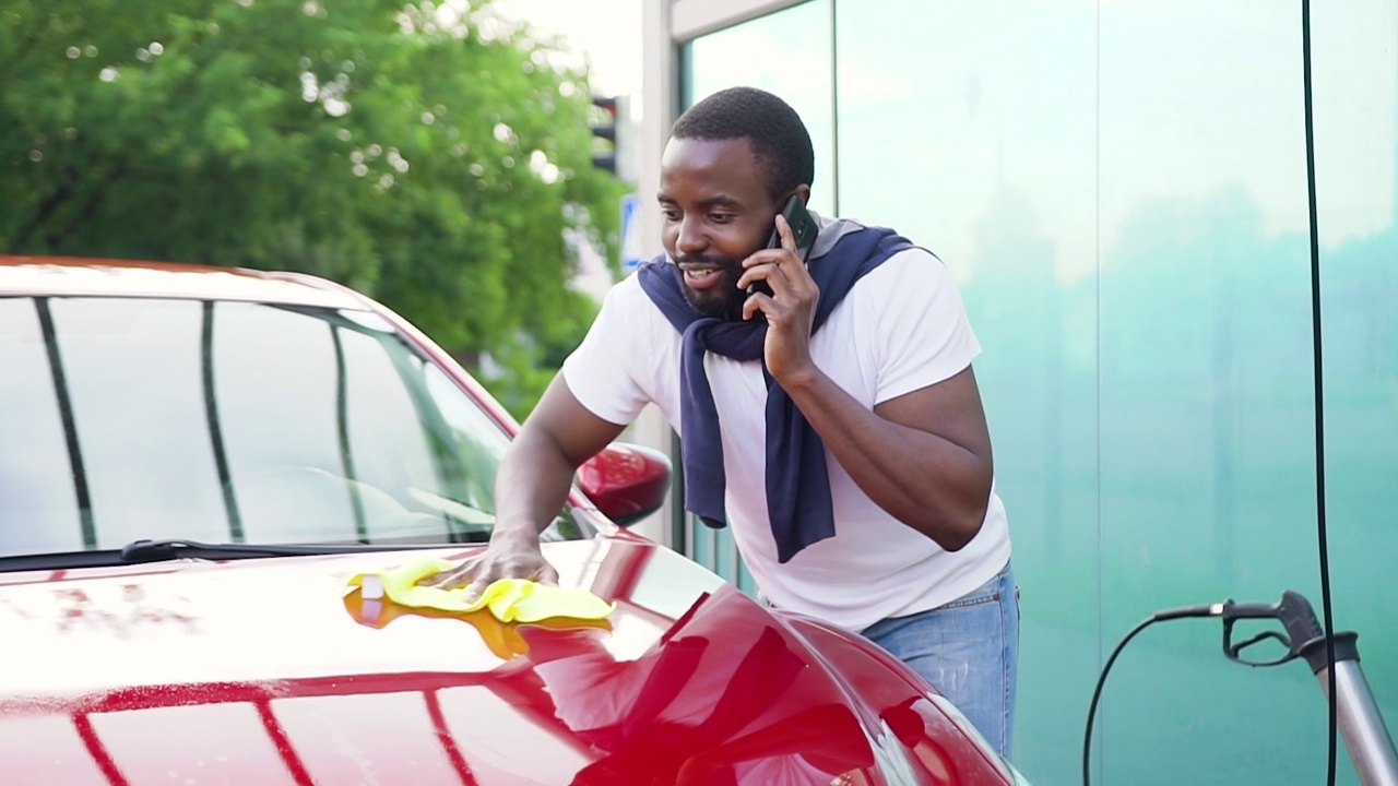 有吸引力的微笑年轻的蓄着胡须的非洲裔美国人有电话交谈，而擦干自己的红色汽车洗涤后洗车