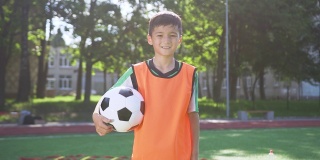 英俊疲惫的亚洲少年足球穿着站在球场与球之间的城市公园树木和严肃的脸看着相机